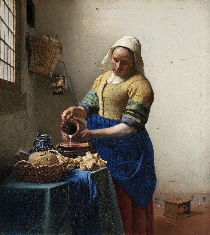 A maid in a yellow bodice and blue apron pours milk from a jug into a bowl beside a window; bread on the table, a small foot warmer on the floor.